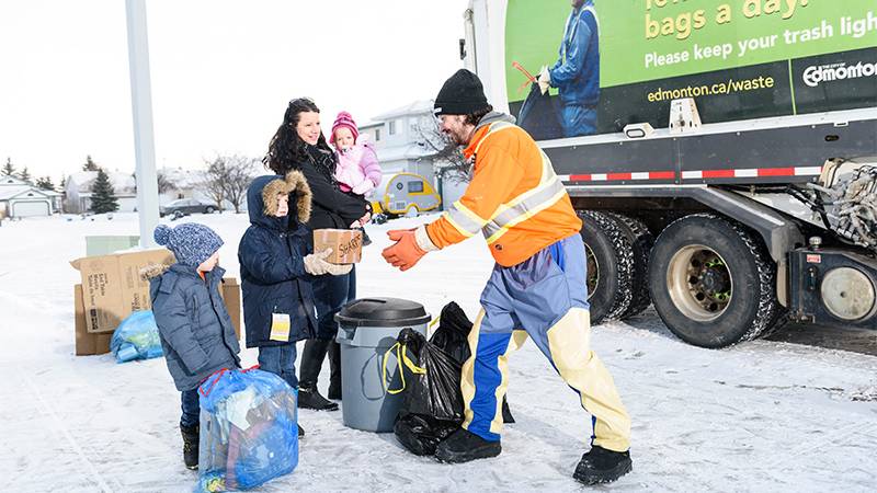 A waste collector picking up household garbage during winter while a family looks on.