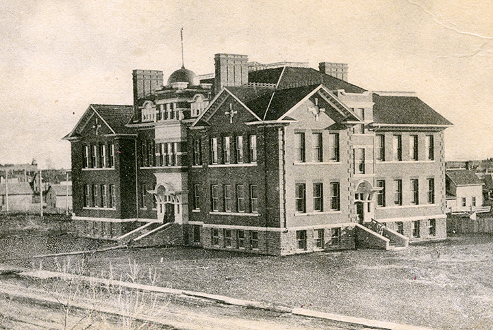 Black and white photo. A multi-story school building. Two entrances with stairs are visible. Multiple windows line the outside of the building at regular intervals.