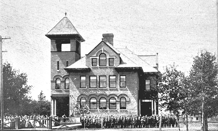 Black and white photo. A large multi-story building with a group of people, most likely students and teachers, standing in front of it.