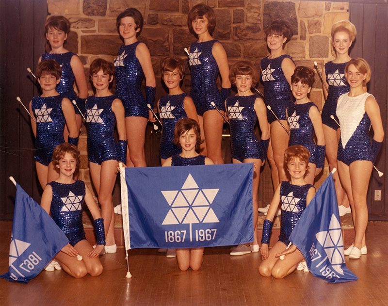 Colour photo of the Klondike Centennial Majorettes, in costume. In front of them is a flag featuring the official Centennial symbol.