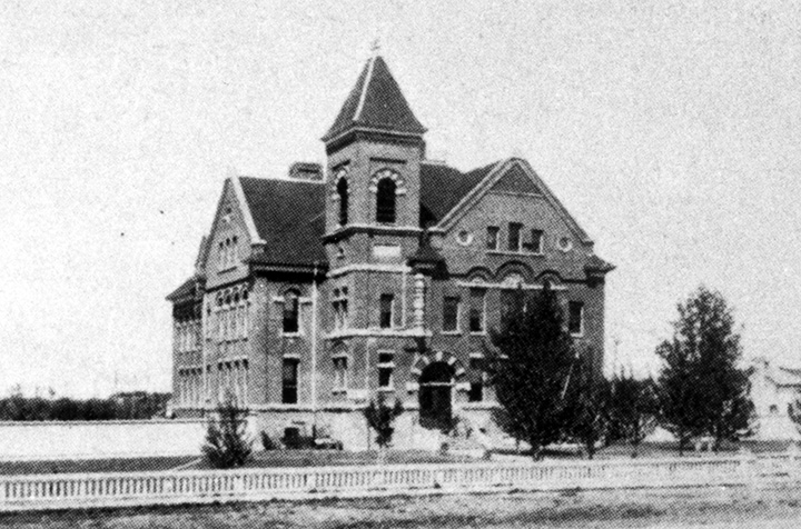 Black and white photo. A large multi-story building with a small white fence in front of it.