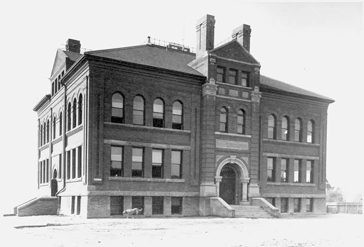 Black and white photo. A large multi-story building. Two entrance or exit doors are visible, each with a set of stairs leading up to it. A dog is walking in front of the building.