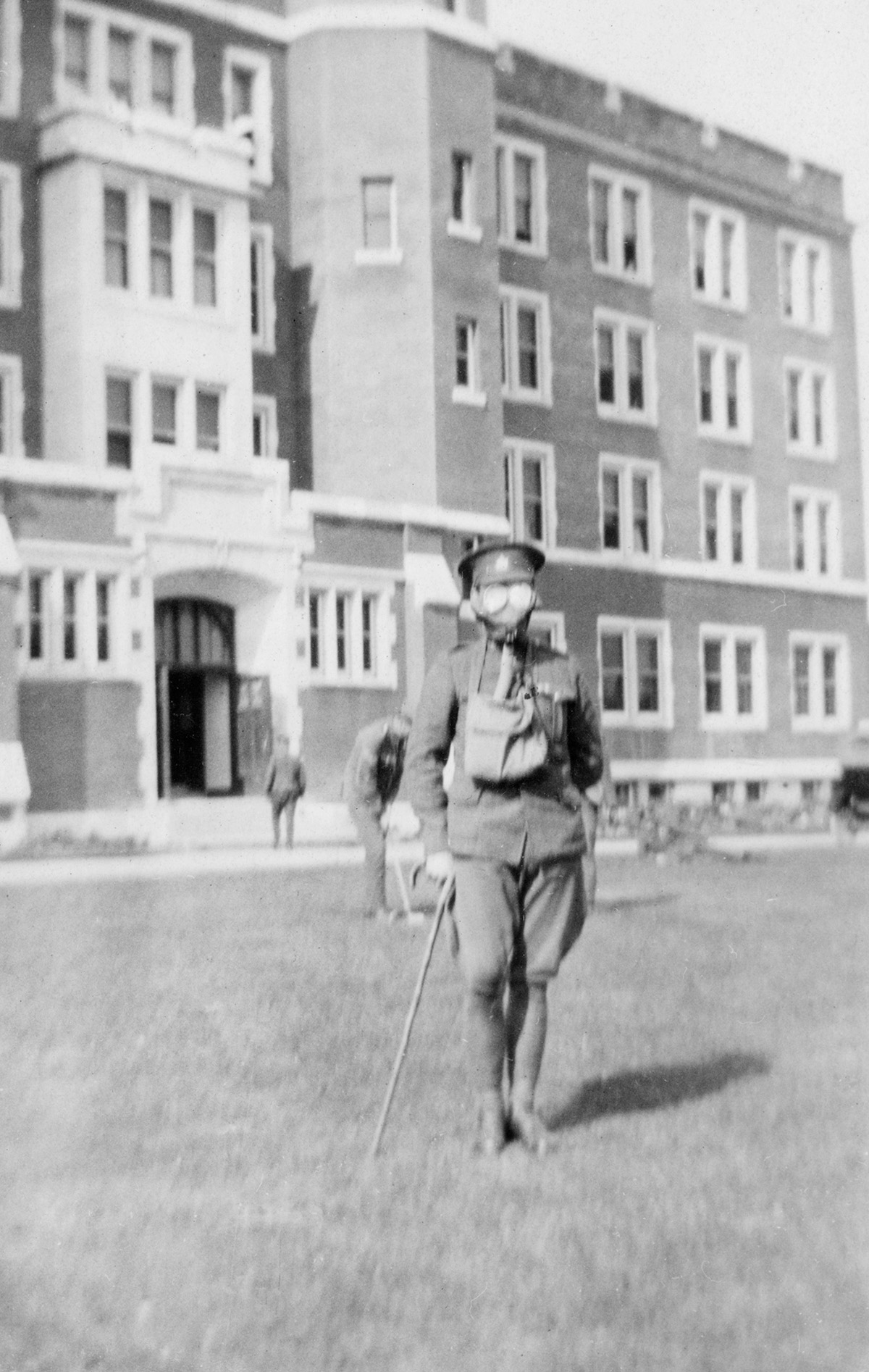Black and white photo of a soldier in a gas mask. He is leaning on a cane in front of Alberta College South Veterans' Hospital.