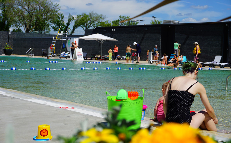 A woman and a child sitting on the edge of Borden Natural Swimming Pool. Groups of people are visible in the background, on the other side of the pool.