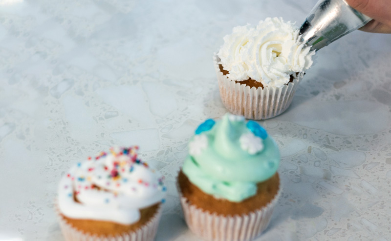 Close-up of icing being piped onto a cupcake. Two already-decorated cupcakes sit in the foreground.