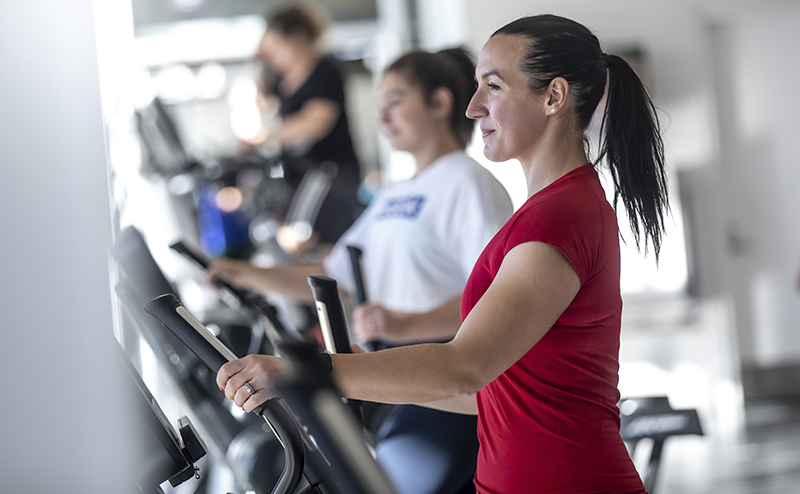 A woman in a red shirt working out on a step machine. Other people are working out on machines in the background, but the woman is the only one in focus.