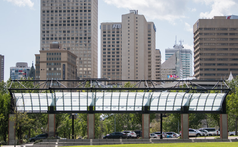 Photo of the Shumka Stage at Louise McKinney Riverfront Park. The stage is a raised platform with steps, covered by a translucent awning overhead. City skyscrapers are visible in the background.