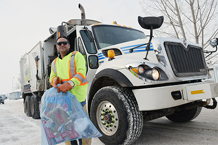 Waste collector holding a blue recycling bag in front of a garbage truck.