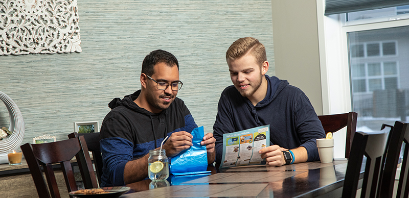 Two men at a kitchen table. One is unfolding a blue bag while the other examines a "What goes where" waste document