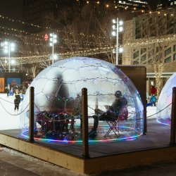 A group of people sitting in a geodescent warming dome in front of the City Hall skating rink