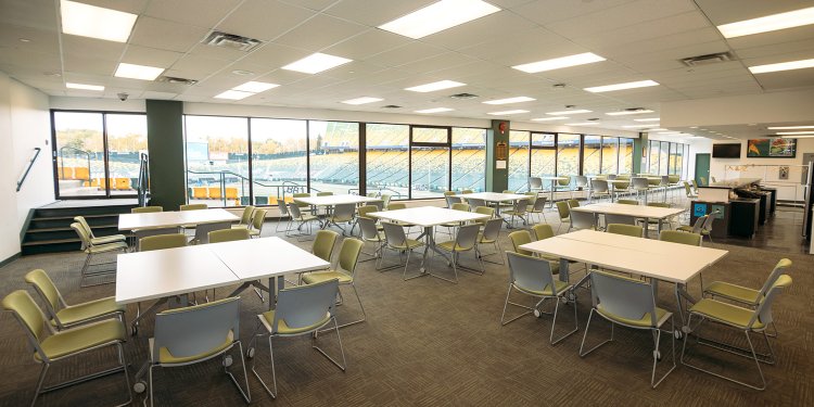 A large room containing paired rectangular tables, with 8 chairs at each set of tables. In the background, round tables with 2 chairs each are also visible. A window at one end of the room overlooks the field at Commonwealth Stadium.