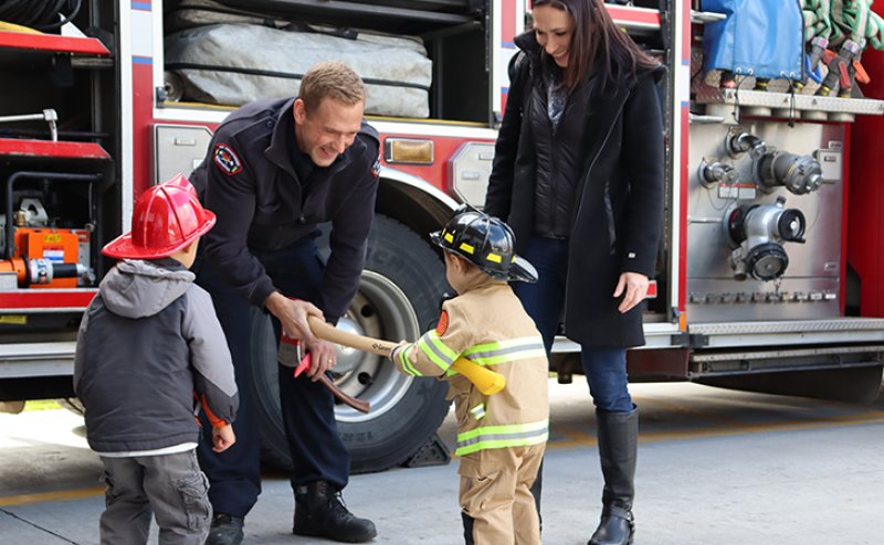 An adult and two kids dressed as firefighters meet a firefighter. The firefighter hands one of the kids a fire axe.