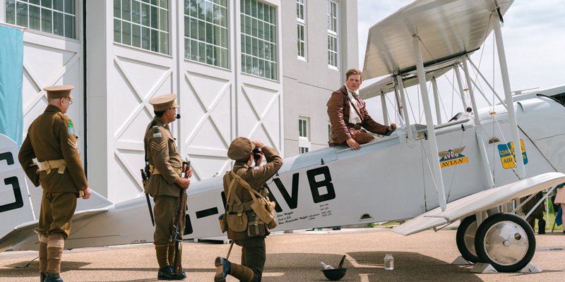 A person sits in an old style plane, while people in period military outfits salute him.
